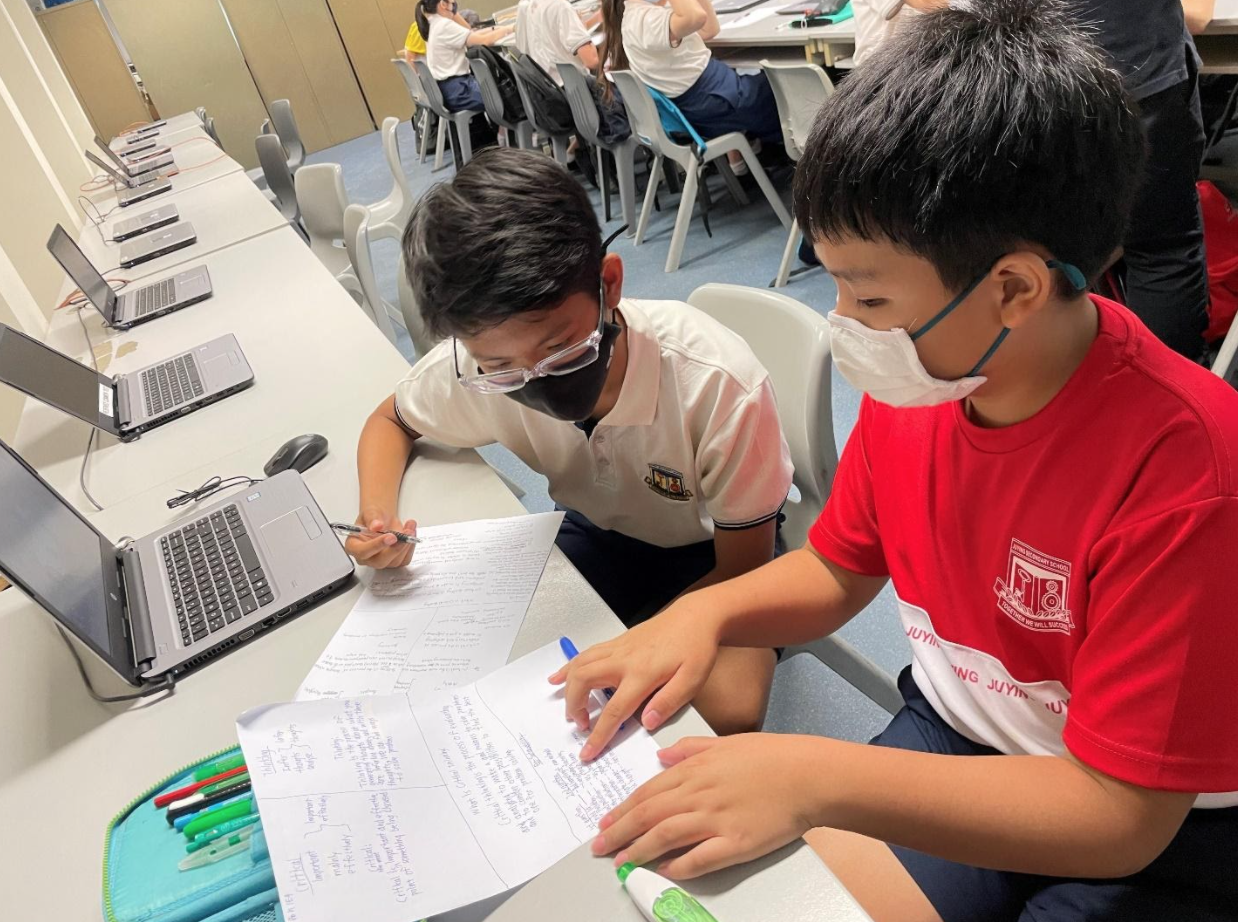 Two masked schoolboys collaborate over handwritten papers at a desk with laptops in a classroom.